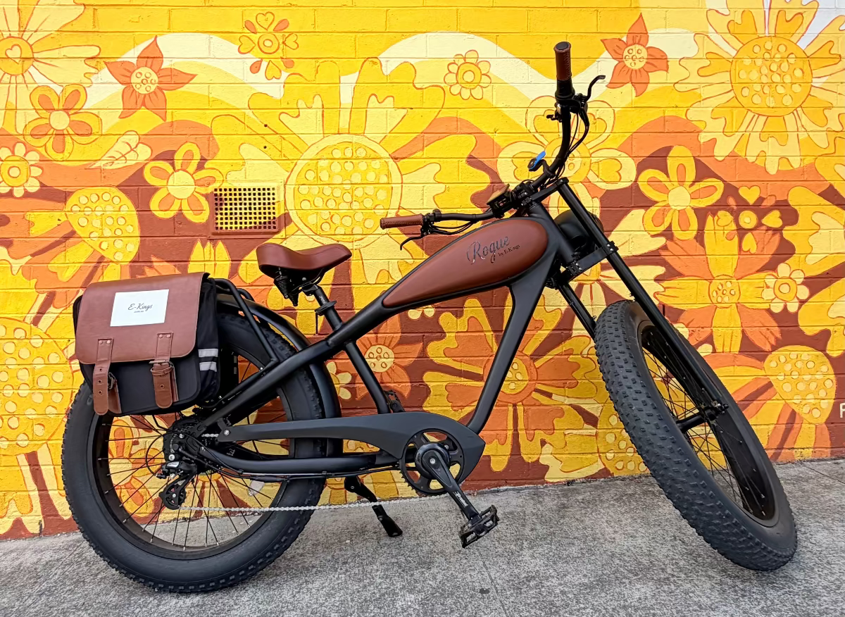Rogue electric fat tire bike with a brown fuel tank and brown saddle, parked in front of a bright yellow and orange floral mural wall.